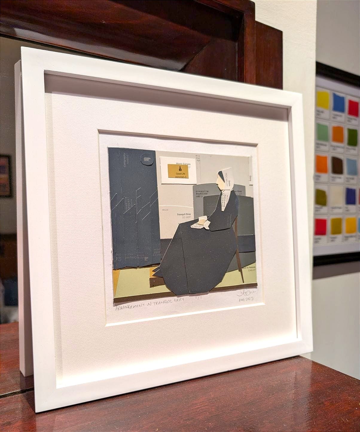 Photo of the piece in a white frame. Background shows a framed collection of paint chips 